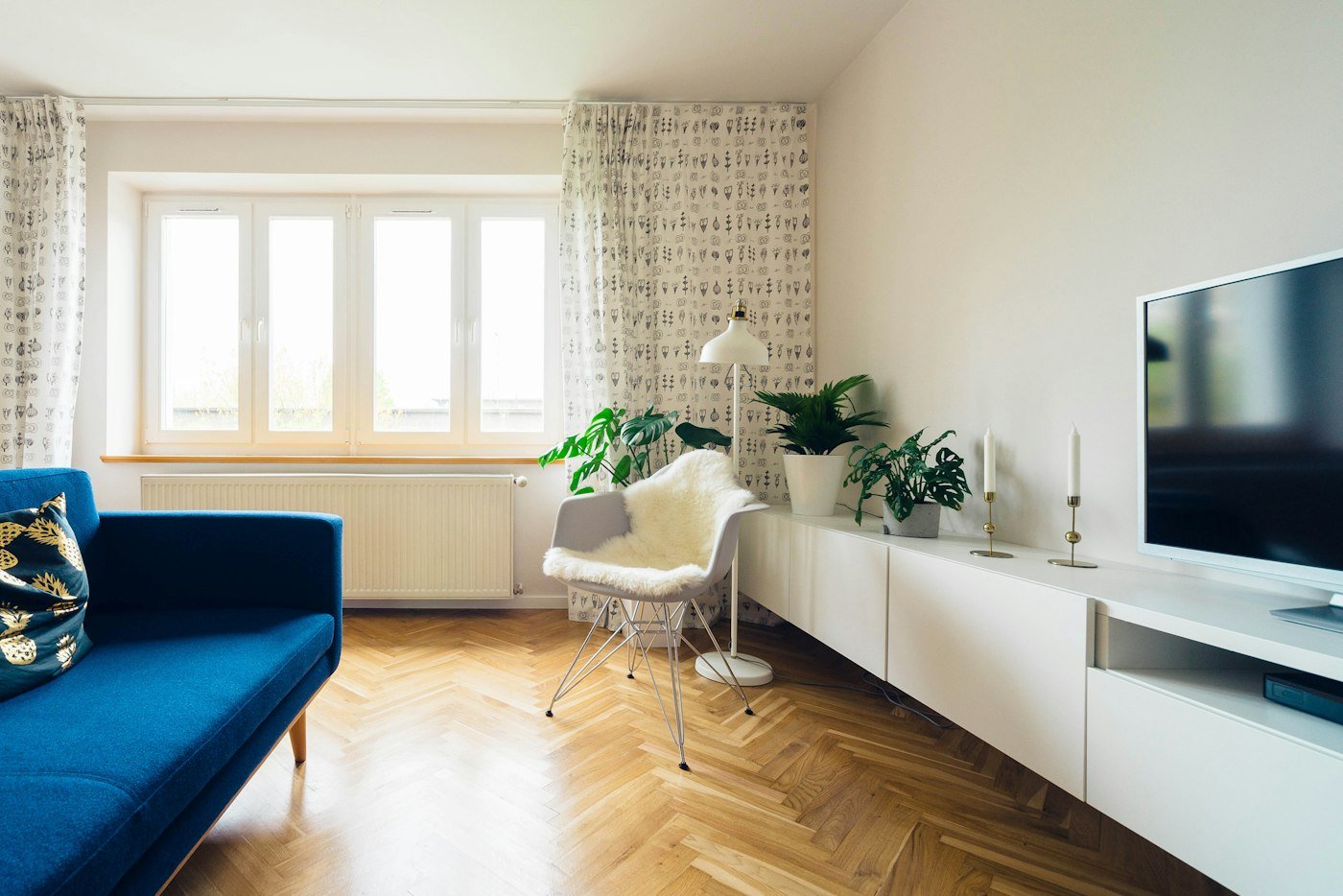 A living room with a blue sofa, herringbone wood floor, houseplants, and a white cupboard under a television