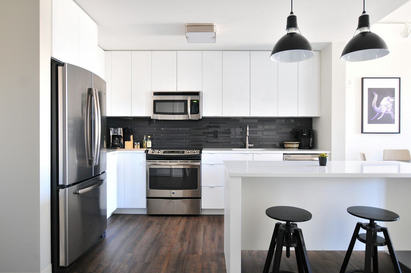 A white kitchen with a steel fridge, black pendant lights, and two stools at an island